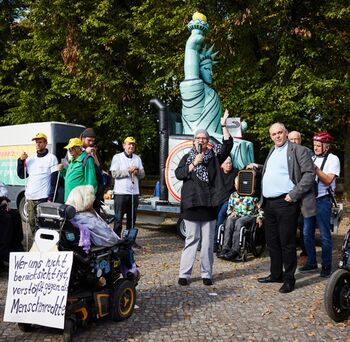 Eine Gruppe von Menschen, manche im Rollstuhl, manche mit Blindenstock, versammelt vor einer 3 Meter hohen aufblasbaren Freiheitsstatue im  Rollstuhl. Vor der Statue steht Martina Puschke, Sie spricht mit erhobenem Arm anfeuernd in ein Mikrofont